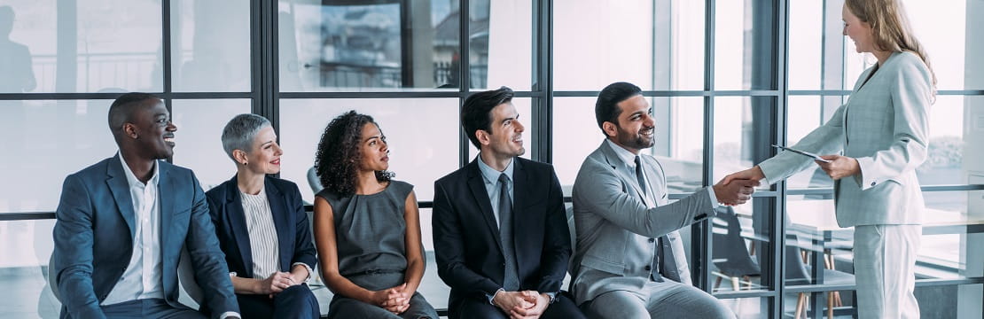 Line of job applicants wait in chairs while one shakes hands with someone.